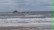 Am Strand sind hohe Wellen und Schaum. Im Hintergrund schüttet ein Schiff Sand auf.